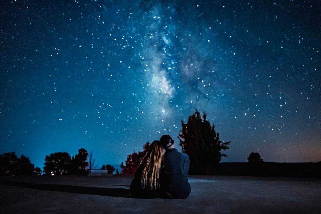 A couple sits together under a starry night sky filled with the Milky Way.